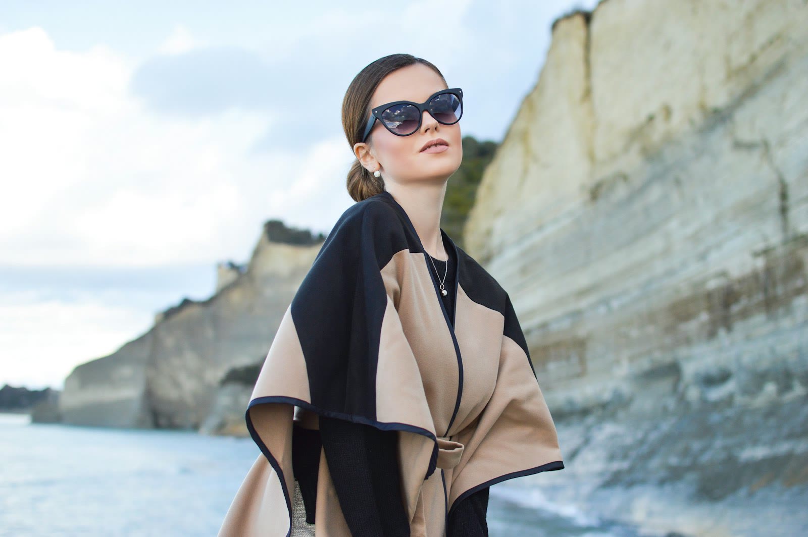 Una donna con occhiali da sole e mantella chiara davanti a una scogliera sul mare